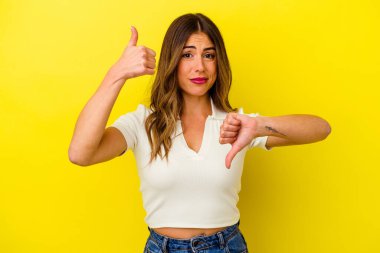 Sarı arka planda izole edilmiş genç beyaz kadın baş parmağını kaldırıyor ve baş parmak aşağı, zor seçim konsepti.