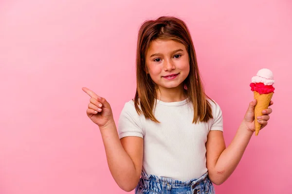 一个拿着冰激凌的高加索小女孩 被粉色背景隔开 微笑着指着旁边 在空旷的空间里展示着一些东西 — 图库照片