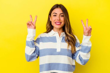 Sarı arka planda izole edilmiş genç beyaz kadın zafer işareti gösteriyor ve gülümsüyor..