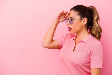 Pembe arka planda izole edilmiş genç beyaz kadın uzaklara bakıyor ve elini alnına koyuyor..