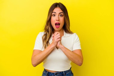 Beyaz tenli genç bir kadın sarı arka planda izole edilmiş şans diliyor, şaşırmış ve ağzını açıp önüne bakıyor..