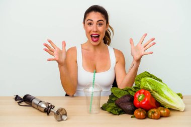 Genç beyaz kadın sebzelerle sağlıklı bir smoothie hazırlıyor zaferi ya da başarıyı kutluyor, şaşırıyor ve şok oluyor..