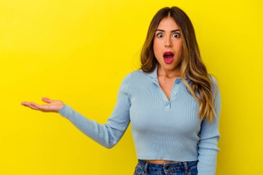 Sarı arka planda izole edilmiş genç beyaz kadın avuç içinin fotokopi alanını elinde tutmaktan etkilenmiş..