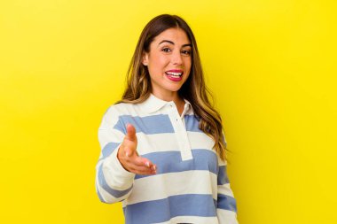 Sarı arka planda izole edilmiş genç beyaz kadın selamlaşma jesti olarak kameraya elini uzatıyor..