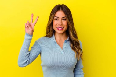 Sarı arka planda izole edilmiş genç beyaz kadın zafer işareti gösteriyor ve gülümsüyor..