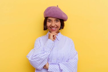 Sarı arka planda izole edilmiş bereli, mutlu ve kendinden emin gülümseyen, elleriyle çenesine dokunan genç bir ırk kadını..