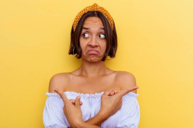 Sarı arka planda izole edilmiş genç melez kadın iki seçenekten birini seçmeye çalışıyor..