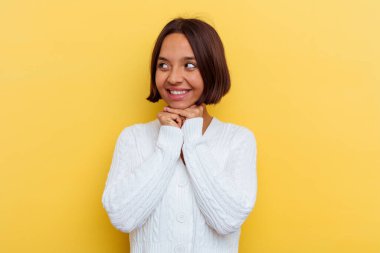 Sarı arka planda izole edilmiş genç melez kadın ellerini çenesinin altında tutuyor, mutlu bir şekilde yana bakıyor..