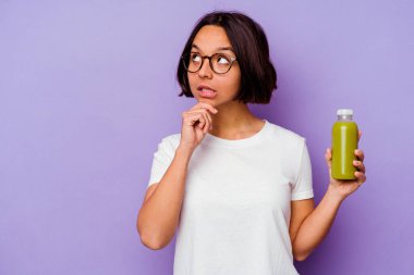 Mor arka planda izole edilmiş sağlıklı bir smoothie tutan genç melez kadın şüpheli ve şüpheci bir ifadeyle yana bakıyor..