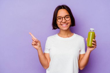 Mor arka planda izole edilmiş sağlıklı bir smoothie tutan genç bir ırk kadını gülümsüyor ve boş alanda bir şeyler gösteriyor..