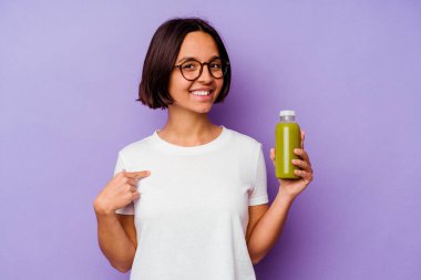 Mor arka planda izole edilmiş sağlıklı bir smoothie tutan genç bir ırk kadını gömlek fotokopi alanını işaret ediyor, gururlu ve kendinden emin.