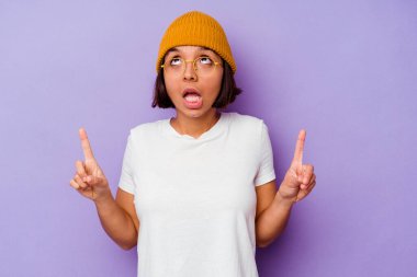 Mor arka plana izole edilmiş yün şapka takan genç bir ırk kadını ağzı açık, üst tarafı işaret ediyor..