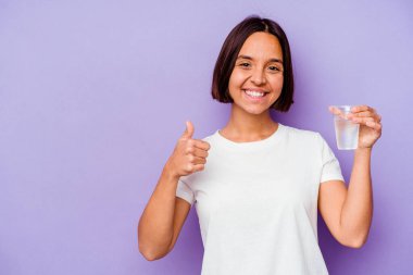 Mor arka planda izole edilmiş bir bardak su tutan melez ırk gülümsüyor ve başparmağını kaldırıyor.