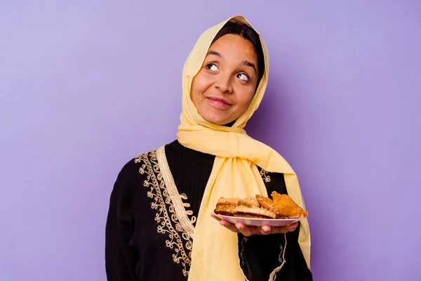 Young Moroccan woman holding a Arabian sweets isolated on purple background dreaming of achieving goals and purposes