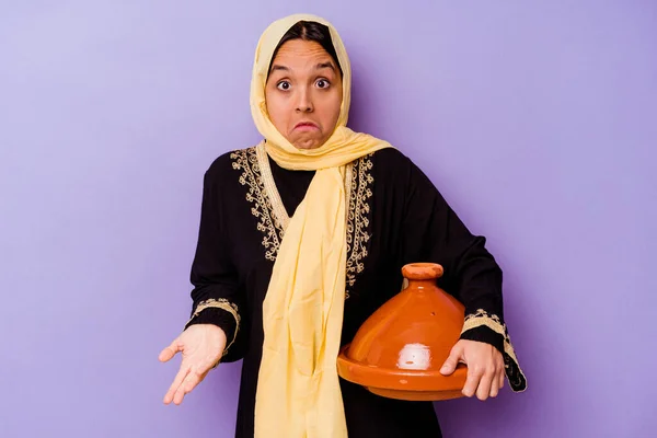 Young Moroccan woman holding a tajine isolated on purple background shrugs shoulders and open eyes confused.