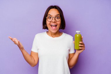 Mor arka planda izole edilmiş sağlıklı bir smoothie tutan genç melez kadın hoş bir sürprizle karşılaşıyor, heyecanlı ve el kaldırıyor..