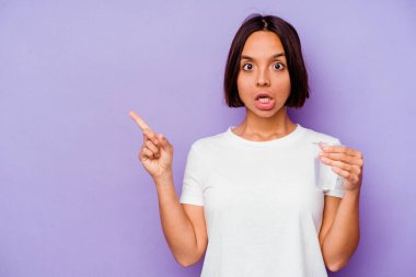 Mor arka planda izole edilmiş bir bardak su tutan genç melez ırk yan tarafı işaret ediyor.