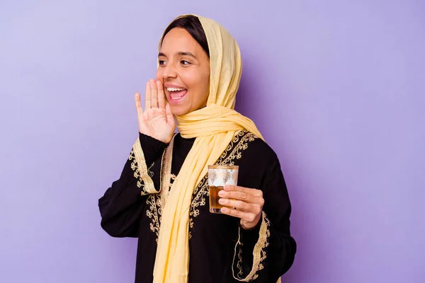 若いモロッコ人女性が紫色の背景の叫びに隔離されたお茶のグラスを持ち 口を開けた近くに手のひらを保持 — ストック写真