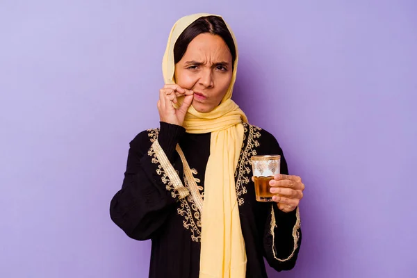 Young Moroccan woman holding a glass of tea isolated on purple background with fingers on lips keeping a secret.