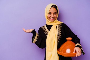 Young Moroccan woman holding a tajine isolated on purple background showing a copy space on a palm and holding another hand on waist. clipart