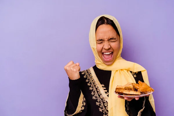 Young Moroccan woman holding a Arabian sweets isolated on purple background raising fist after a victory, winner concept.