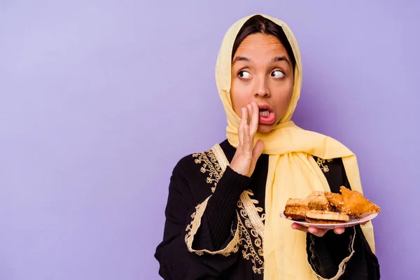 Young Moroccan woman holding a Arabian sweets isolated on purple background is saying a secret hot braking news and looking aside