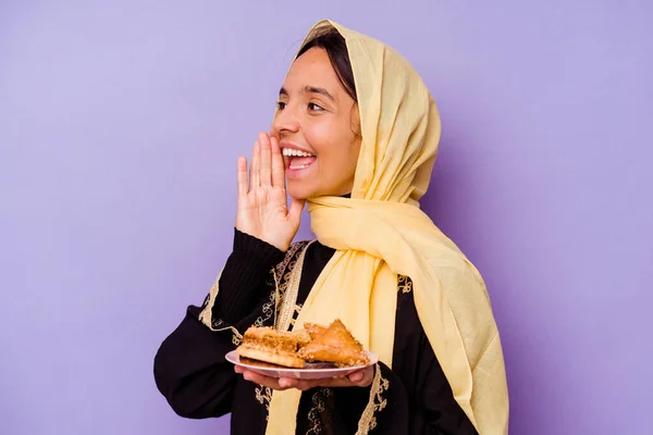若いモロッコ人女性が紫色の背景に隔離されたアラブのお菓子を持って叫びと口を開けた近くに手のひらを保持 — ストック写真