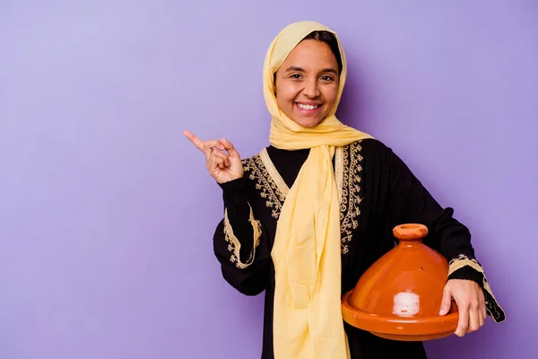 Young Moroccan woman holding a tajine isolated on purple background smiling and pointing aside, showing something at blank space.