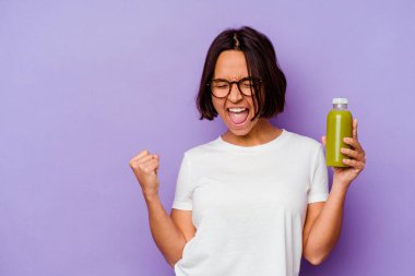 Mor arka planda izole edilmiş sağlıklı bir smoothie tutan genç melez kadın zafer kazandıktan sonra yumruğunu kaldırıyor, kazanan konsept..