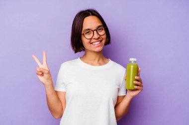 Mor arka planda izole edilmiş sağlıklı bir smoothie tutan genç melez kadın neşeli ve kaygısız parmaklarıyla bir barış sembolü gösteriyor..