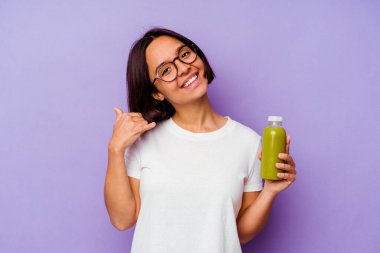 Mor arka planda izole edilmiş sağlıklı bir smoothie tutan genç melez kadın parmaklarıyla bir cep telefonu hareketi gösteriyor..