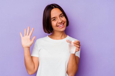 Mor arka planda bir bardak su tutan genç melez ırk gülümseyerek beş numarayı parmaklarıyla gösteriyor..