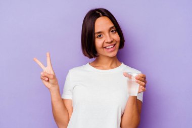 Mor arka planda izole edilmiş bir bardak su tutan genç melez ırk neşeli ve kaygısız parmaklarıyla barış sembolü gösteriyor..