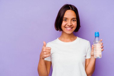 Mor arka planda izole edilmiş su şişesini tutan genç melez ırk gülümsüyor ve başparmağını kaldırıyor.