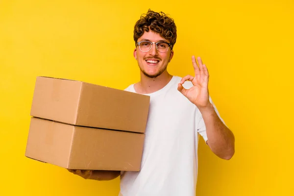 黄色の背景に孤立した段ボール箱を持っている若い白人男性陽気で自信のあるショーOkジェスチャー — ストック写真