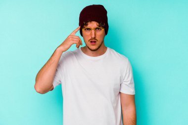 Mavi arka planda yünlü şapka takan genç beyaz adam işaret parmağıyla yaptığı hayal kırıklığı jestini gösteriyor..