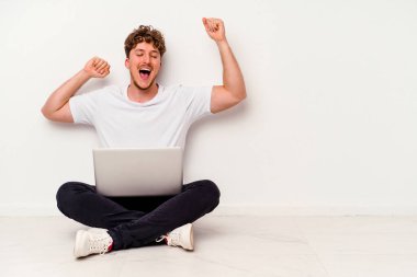 Beyaz arka planda dizüstü bilgisayarı olan genç beyaz bir adam özel bir günü kutluyor, atlıyor ve enerjiyle kollarını kaldırıyor..