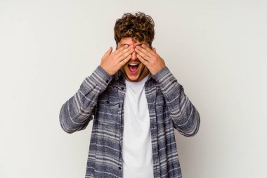 Beyaz arka planda soyutlanmış beyaz tenli genç bir adam elleriyle gözlerini kapatıyor, geniş bir gülümsemeyle bir sürpriz bekliyor..