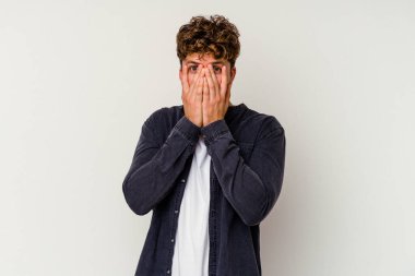 Beyaz arka planda izole edilmiş genç beyaz bir adam parmaklarının arasından korkmuş ve gergin bir şekilde göz kırpıyor..