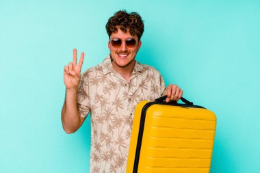 Genç gezgin, mavi arka planda sarı bir bavul tutuyor. İki numarayı parmaklarıyla gösteriyor..