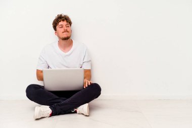 Beyaz arka planda izole edilmiş dizüstü bilgisayarla yerde oturan beyaz tenli genç adam karnına dokunuyor, hafifçe gülümsüyor, yemek yiyor ve tatmin oluyor..