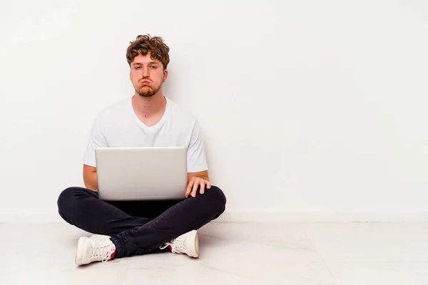 Young caucasian man sitting on the floor holding on laptop isolated on white background blows cheeks, has tired expression. Facial expression concept.