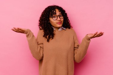 Genç melez kadın pembe arka planda izole edilmiş şüphe içinde ve sorgulama sırasında omuz silkiyor..