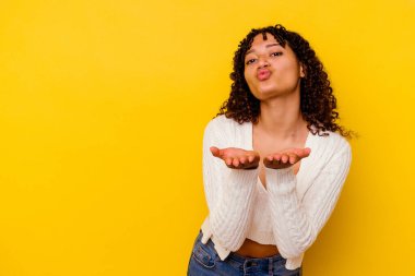 Sarı arka planda izole edilmiş genç melez bir kadın dudaklarını katlıyor ve hava öpücüğü yollamak için avuç içi tutuyor..
