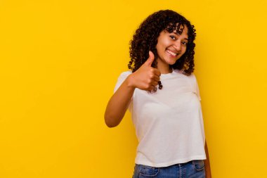 Sarı arka planda izole edilmiş genç melez kadın gülümsüyor ve başparmağını kaldırıyor.