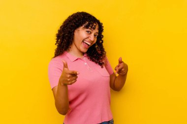Sarı arka planda izole edilmiş genç melez kadın parmaklarıyla ön tarafı işaret ediyor..
