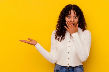 Sarı arka planda izole edilmiş genç melez kadın avuç içinde fotokopi alanı tutuyor, el üstünde tutuyor. Şaşırmış ve mutlu..