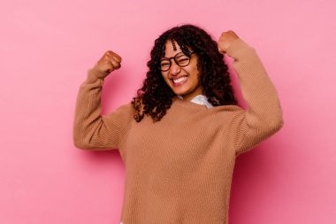 Pembe arka planda soyutlanmış genç melez bir kadın güçlü bir jest yapıyor, kadınsı gücün sembolü.