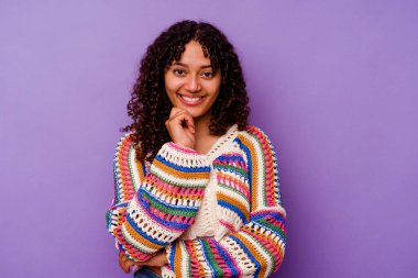Mor arka planda izole edilmiş genç bir ırk kadını mutlu ve kendinden emin gülümsüyor, elleriyle çenesine dokunuyor..