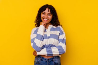 Sarı arka planda izole edilmiş, mutlu ve kendinden emin gülümseyen, elleriyle çenesine dokunan genç bir ırk kadını..